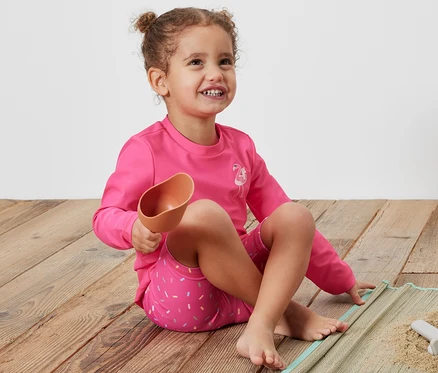 Fillette assise sur une couverture tenant une pelle, portant une combinaison rose pour enfants avec un indice de protection UV 80.