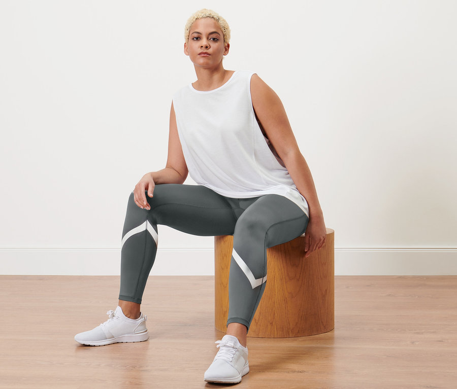 Une femme est assise sur un tabouret en bois et porte un legging de sport gris et 2 débardeurs de sport, gris et blanc.