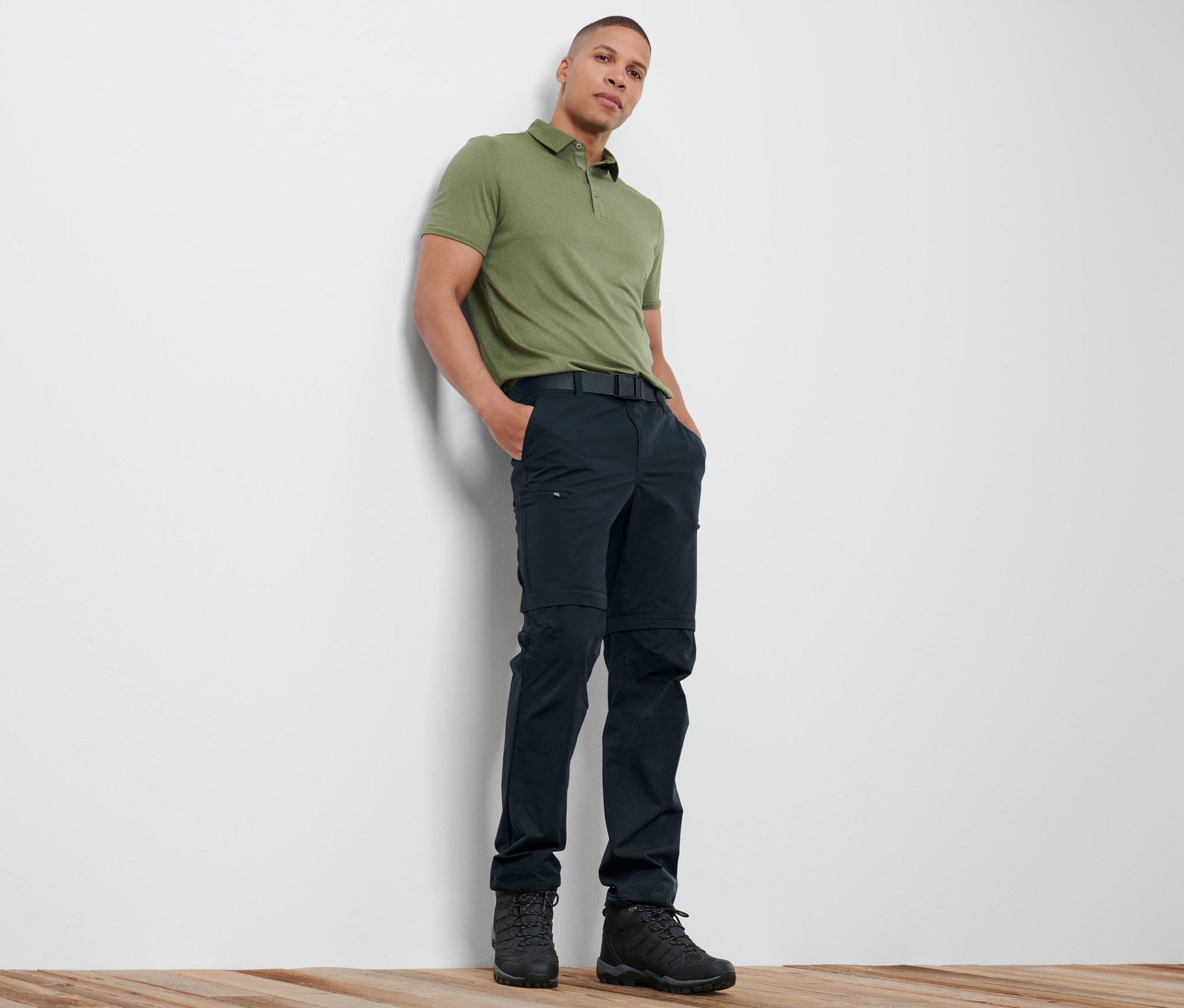 Un homme appuyé contre un mur portant un polo vert et un pantalon fonctionnel 2 en 1 bleu marine.