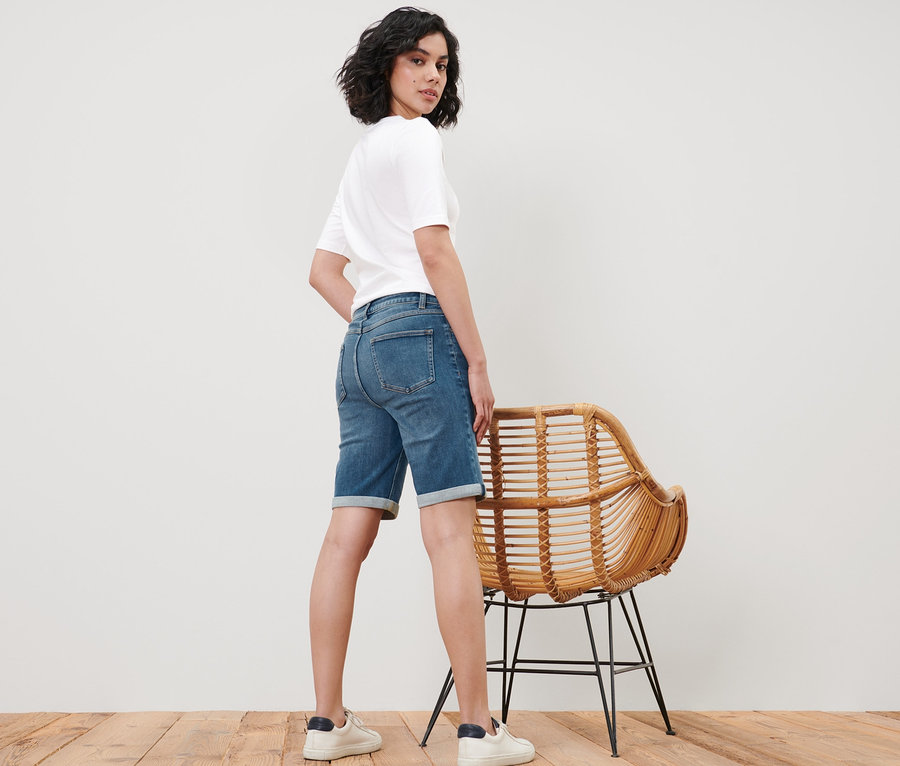Femme posant en bermuda en jean – Fit « Lea » et t-shirt blanc à côté d'une chaise en osier.