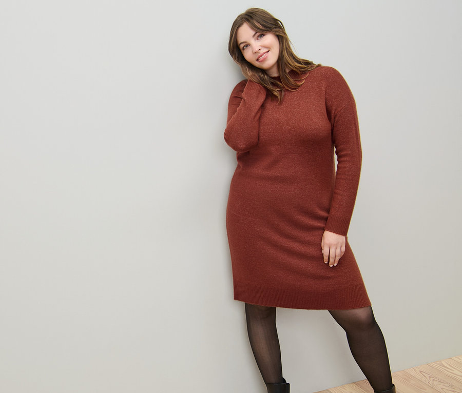 Une femme appuyée contre un mur porte une robe en maille avec teneur en laine et des collants fins 50 DEN en aspect transparent.