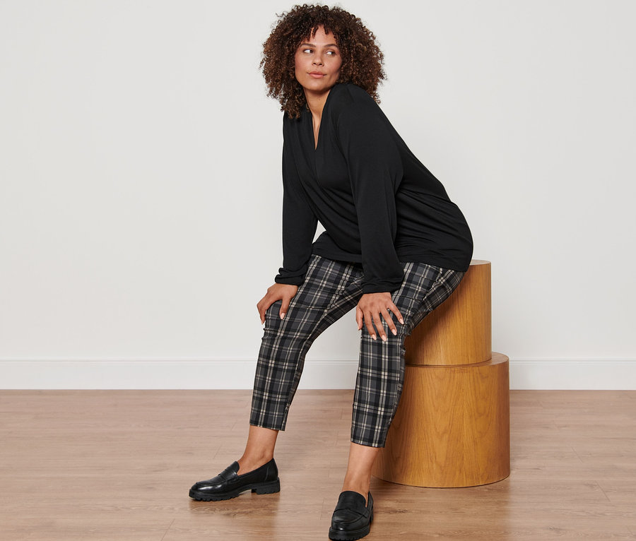 Une femme est assise sur un tabouret en bois et porte un t-shirt manches longues noir avec encolure en V, un pantalon stretch à carreaux et des mocassins en cuir.