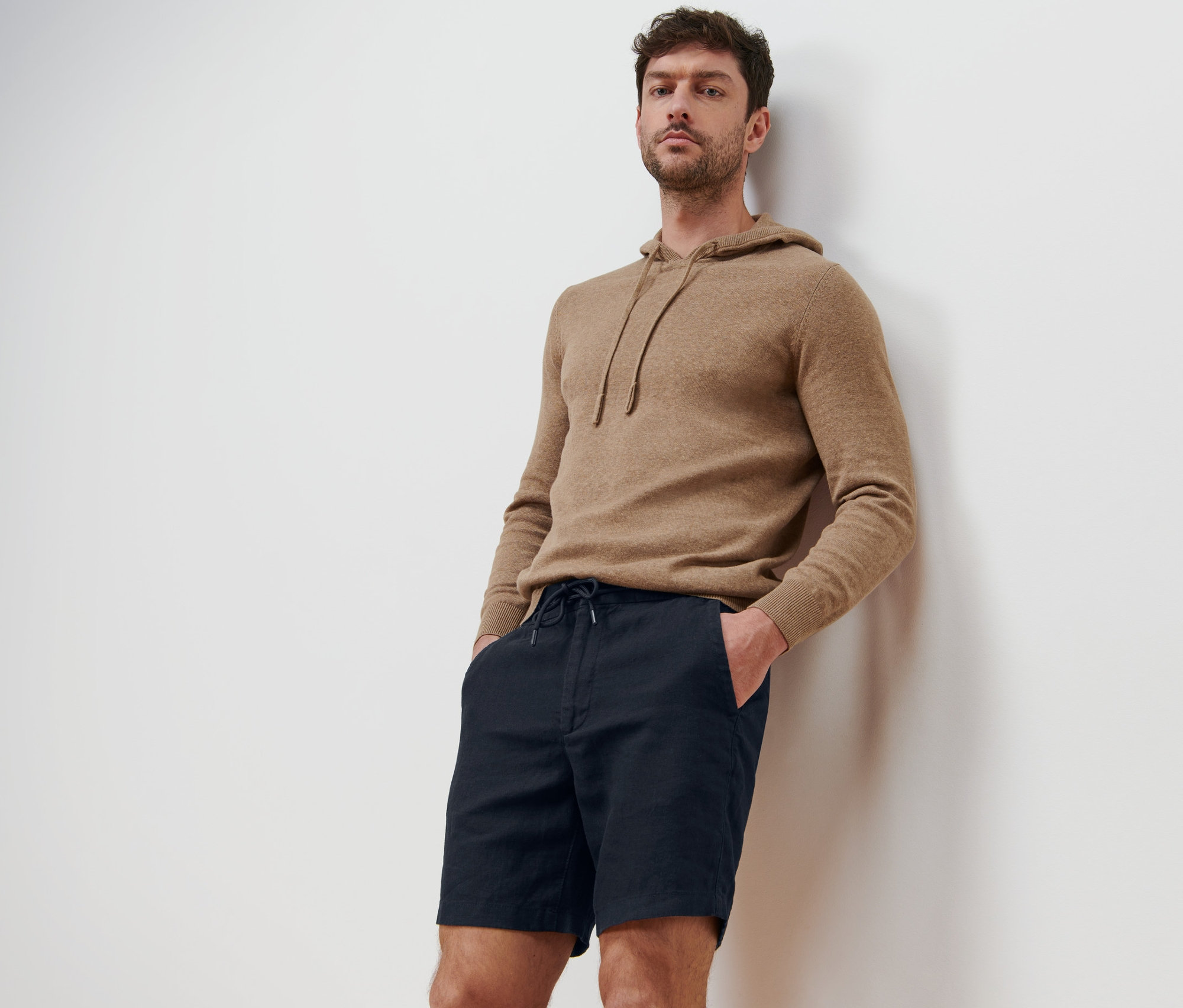 Un homme appuyé contre un mur portant un short avec du lin et un sweat à capuche.