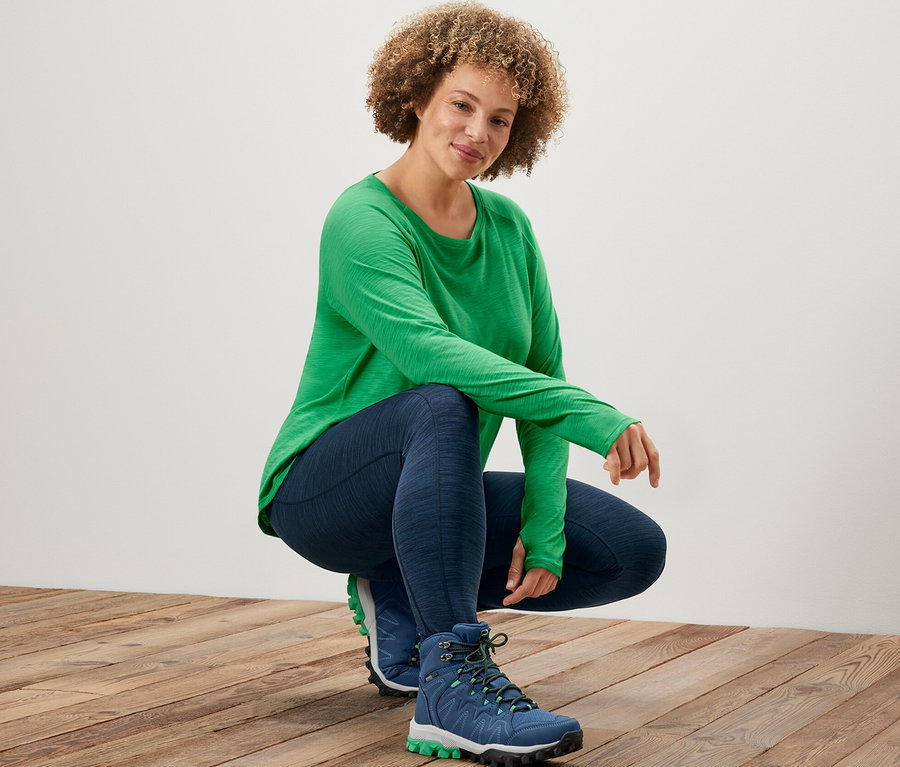 Une femme portant un t-shirt de sport manches longues vert et un legging de sport pose sur un plancher en bois.