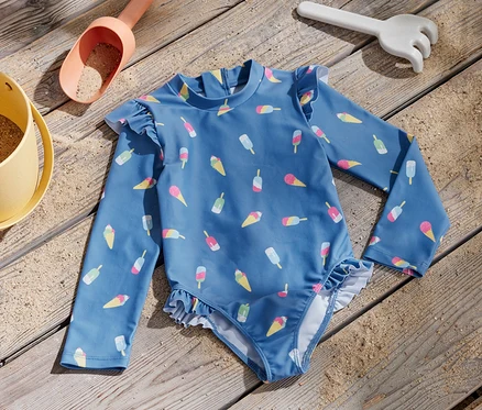 Un maillot de bain bleu pour enfant avec un imprimé de glace et un indice de protection UV 80 est posé sur une planche en bois à côté d'un jouet de sable.