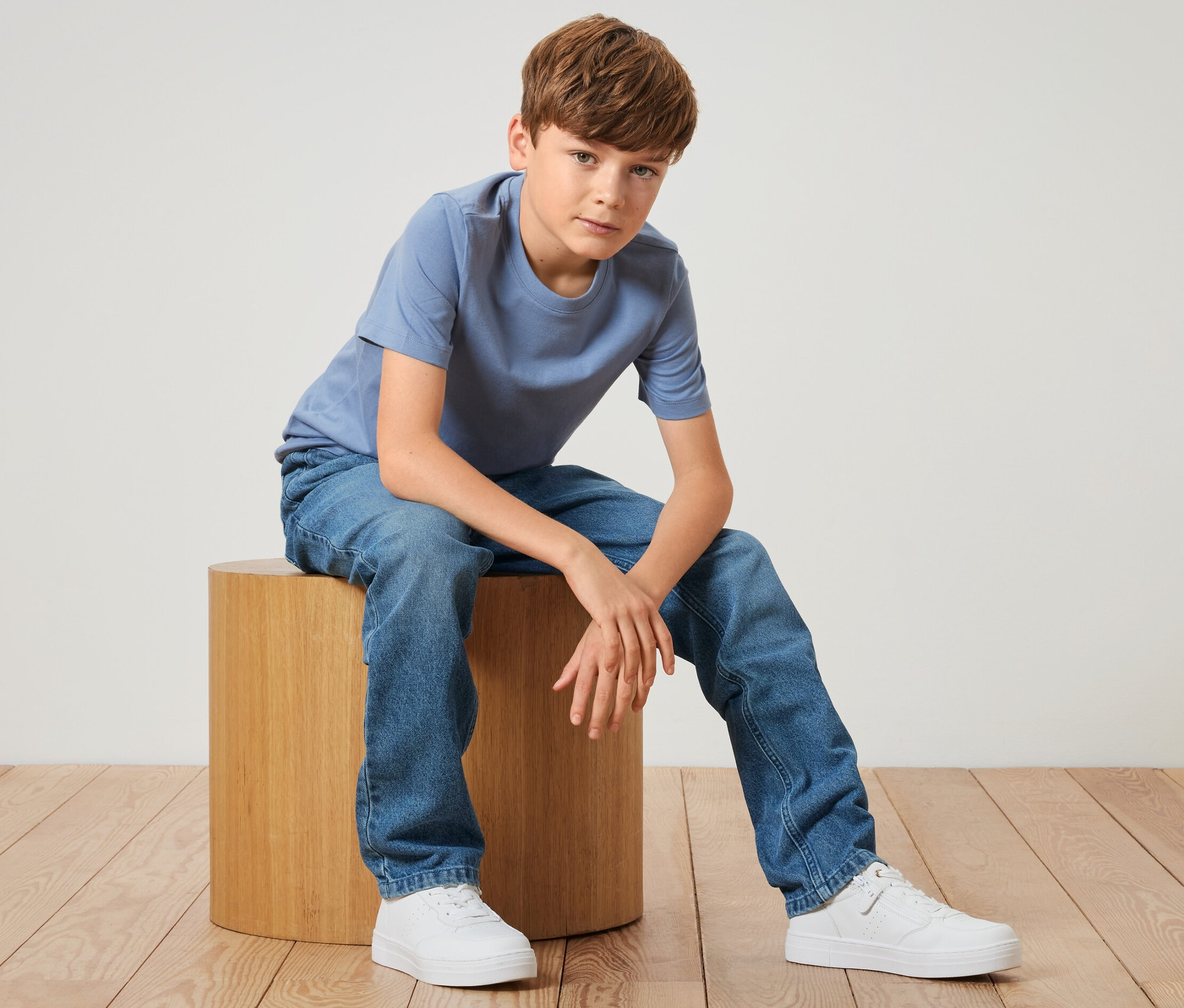 Garçon assis sur un tabouret en bois portant un jean pour enfant – Fit « Jonas » bleu et un t-shirt bleu.