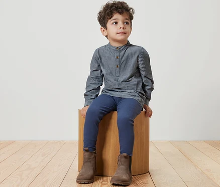 Un garçon est assis sur un tabouret en bois et porte une chemise pour enfant, un legging bleu et des bottes marron.