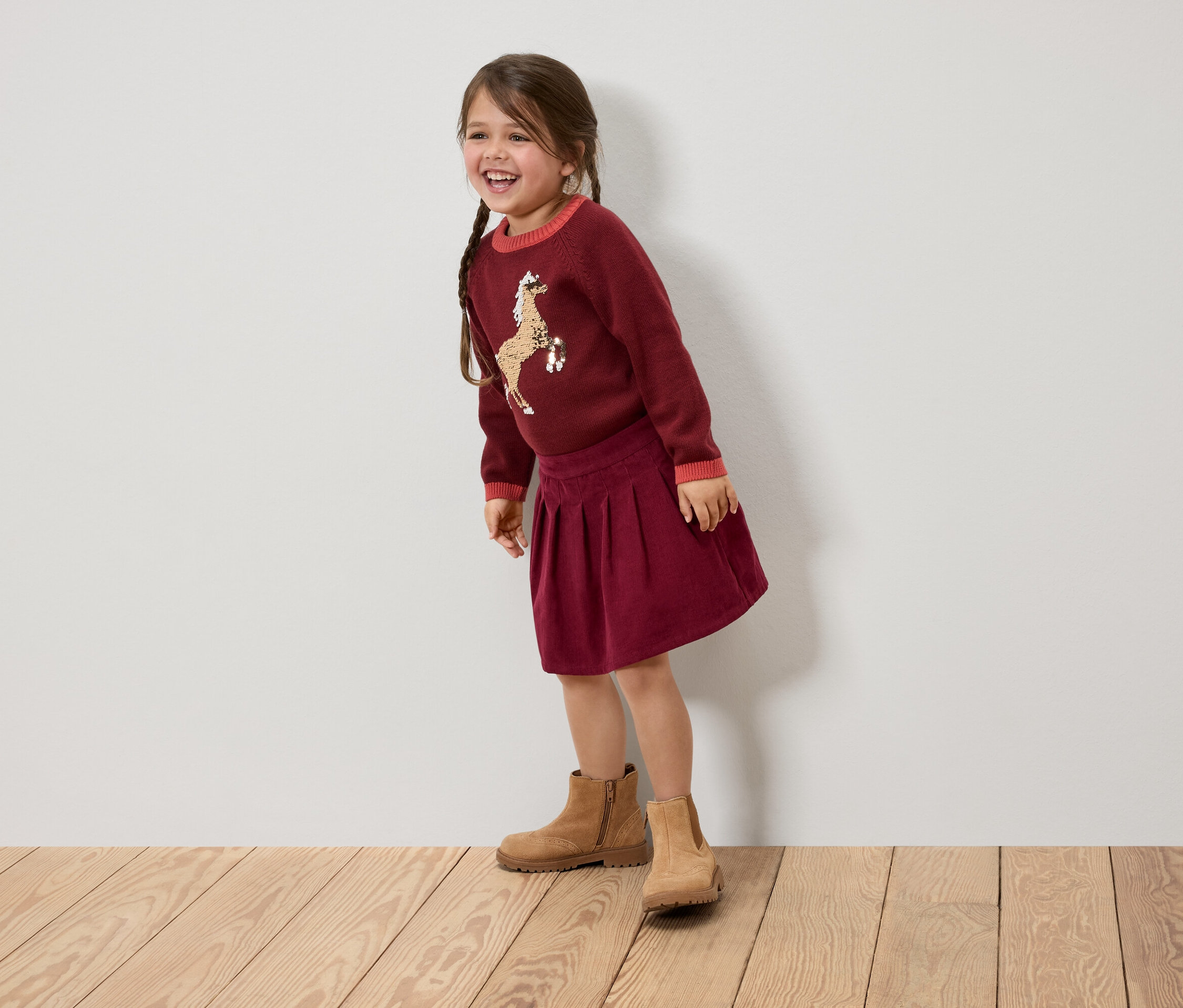 Une fillette souriante avec des tresses porte un pull bordeaux avec un cheval, une jupe et des bottes marrons.