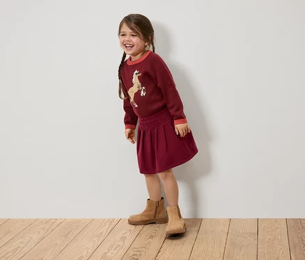 Une fillette souriante avec des tresses porte un pull bordeaux avec un cheval, une jupe et des bottes marrons.