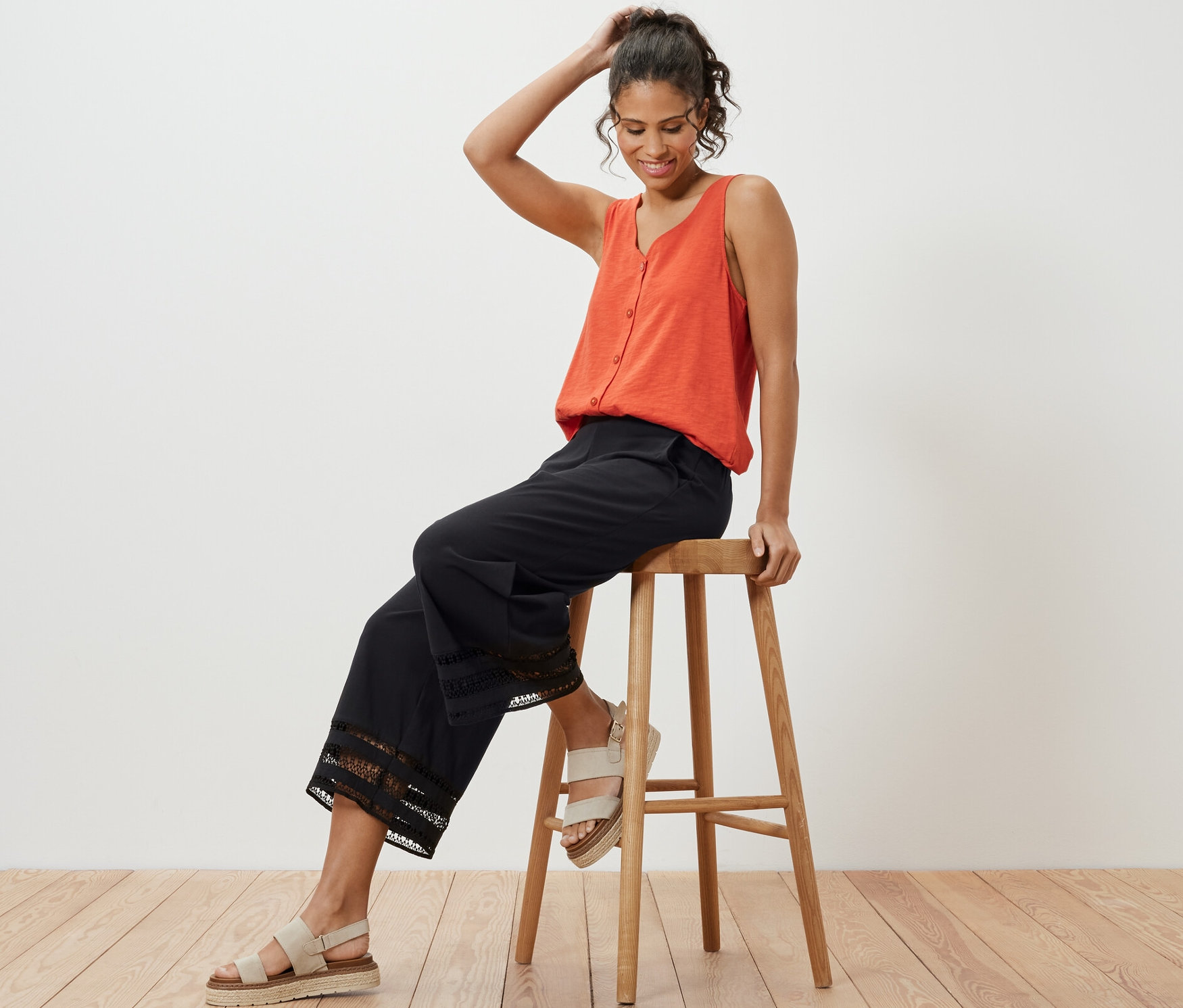 Femme assise sur un tabouret en bois portant un débardeur orange et un pantalon large noir à broderie.