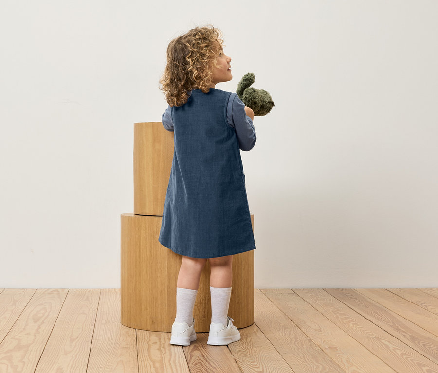 Une fille en robe bleue tient une peluche et se tient à côté de blocs de bois.