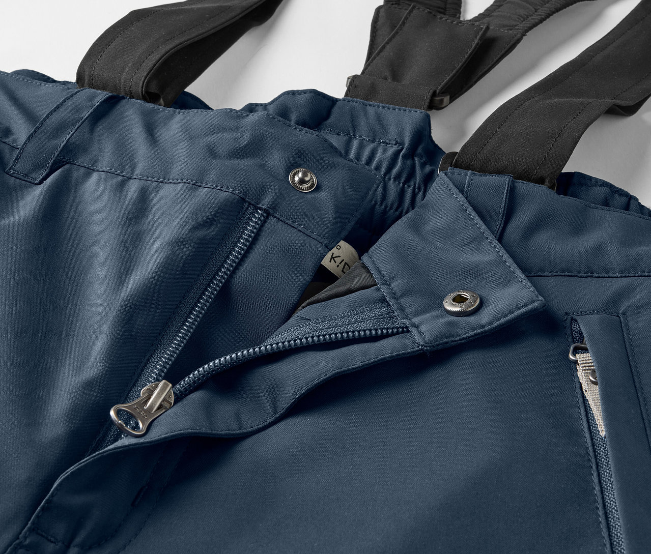 Gros plan sur un pantalon bleu avec des bretelles noires, une fermeture éclair ouverte et des boutons argentés.