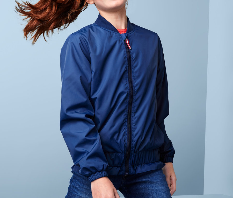 Une fille avec une queue de cheval rousse pose en veste style bomber recyclée et en jean.