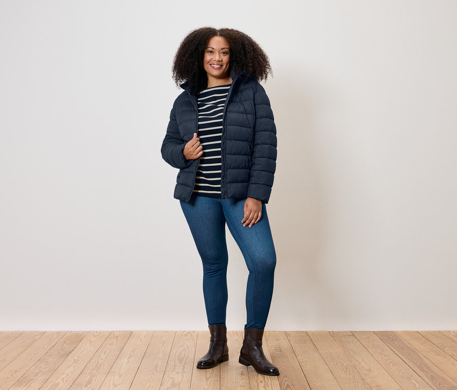 Une femme aux cheveux bouclés se tient debout et porte une veste matelassée bleue, un t-shirt rayé, un jean et des bottes.