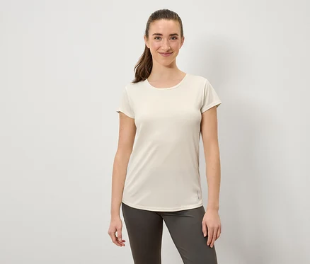 Une femme souriante portant un t-shirt crème et un pantalon gris pose devant un fond blanc.