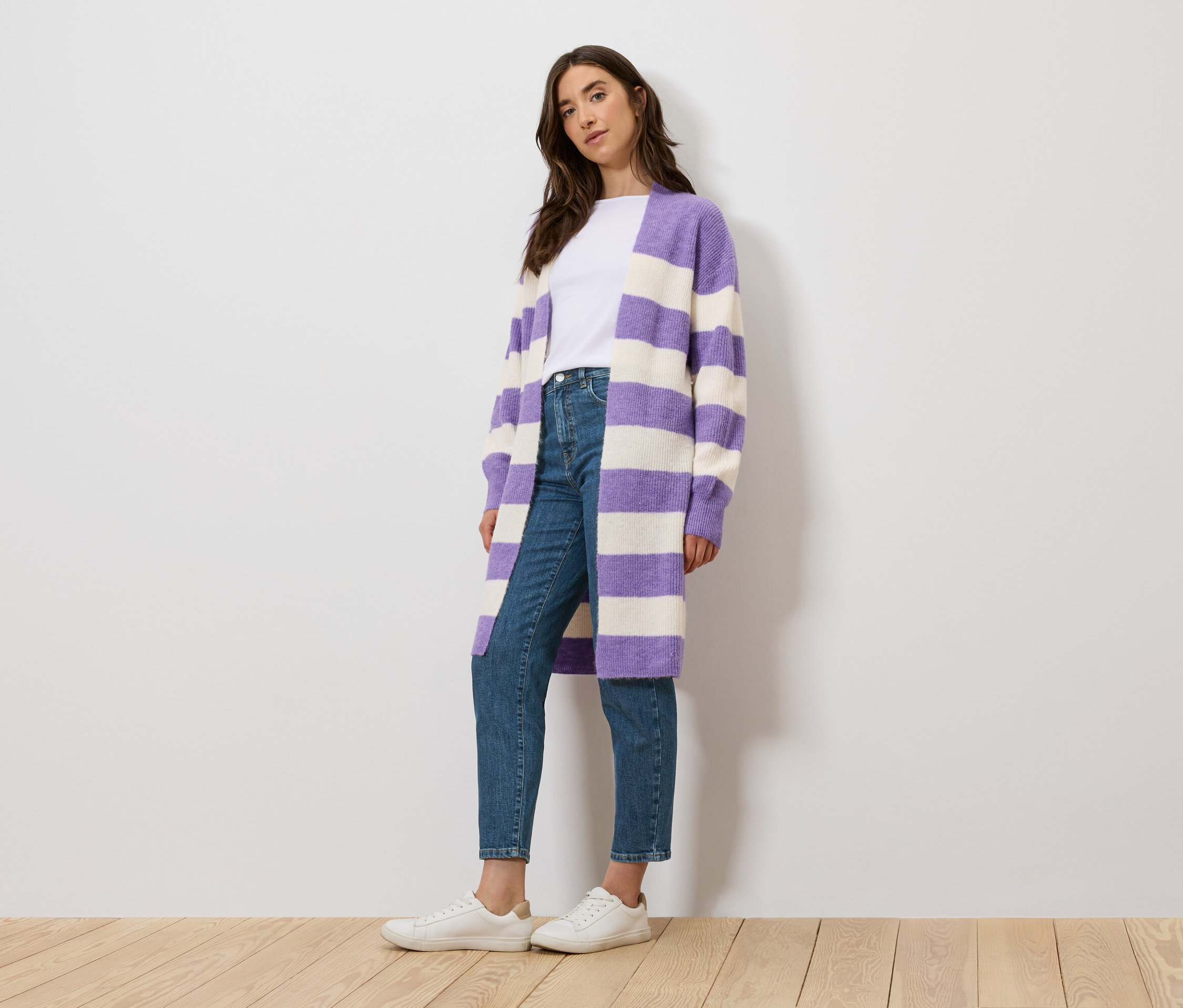 Une femme se tient debout et porte un t-shirt blanc, un jean et un cardigan rayé violet et blanc.