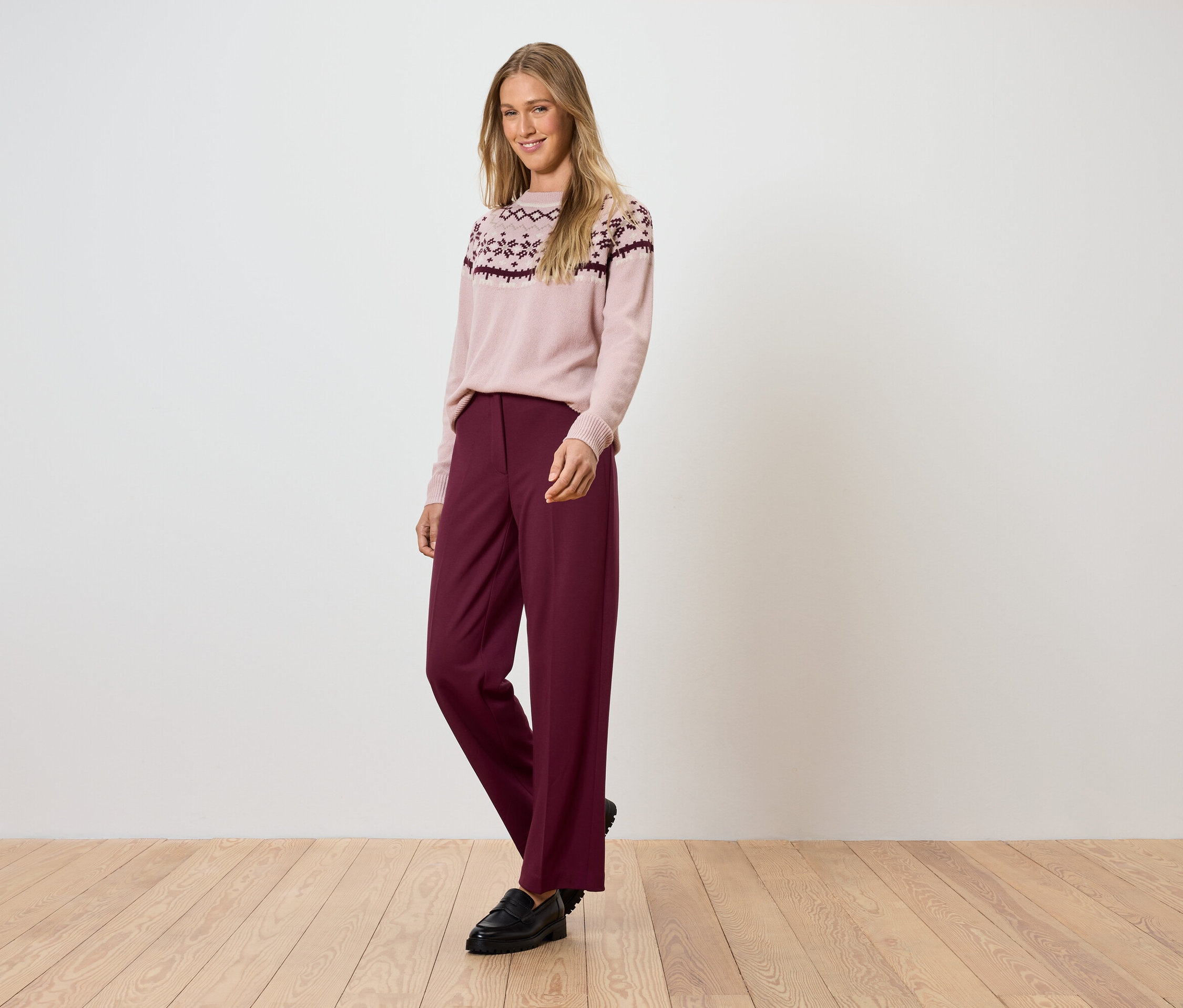 Femme posant dans un pull rose à motifs et un pantalon bordeaux.