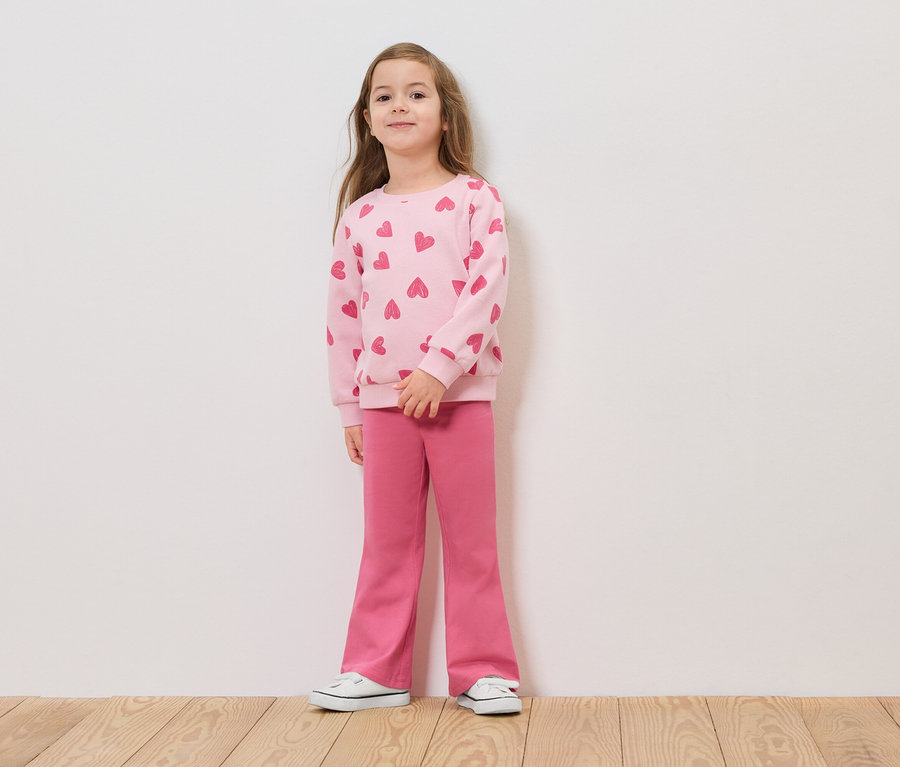 Une fille se tient devant un mur blanc portant un pull rose à motifs de cœurs et un pantalon rose.