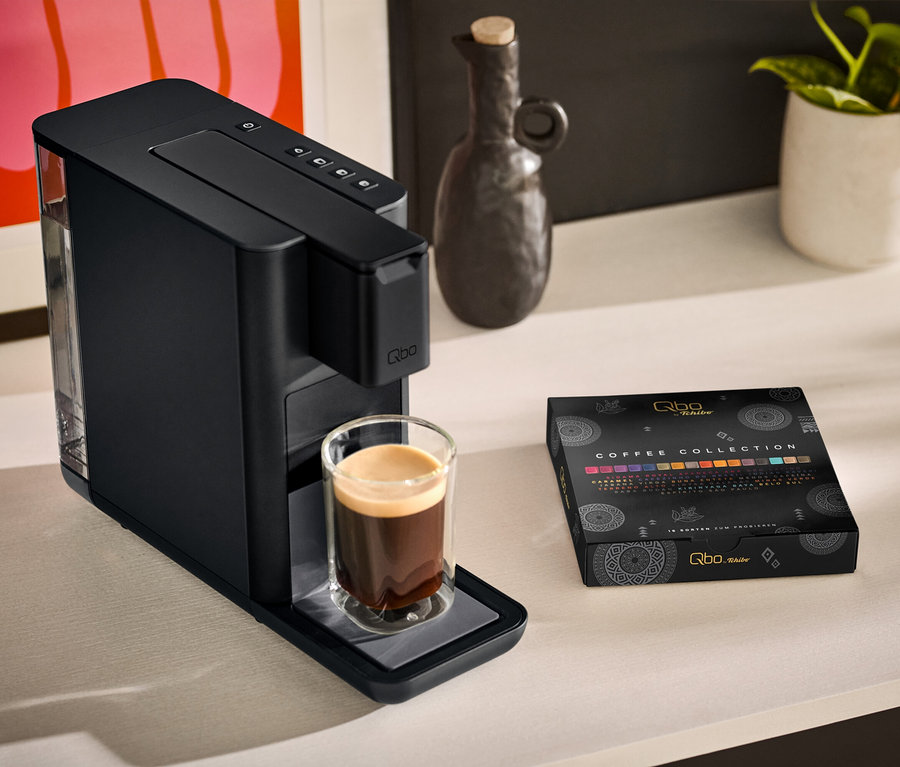 Une machine à café Qbo ICONIC Dark Chrome avec une tasse de café à côté et un paquet de Qbo Collection - 16 capsules sur une table.