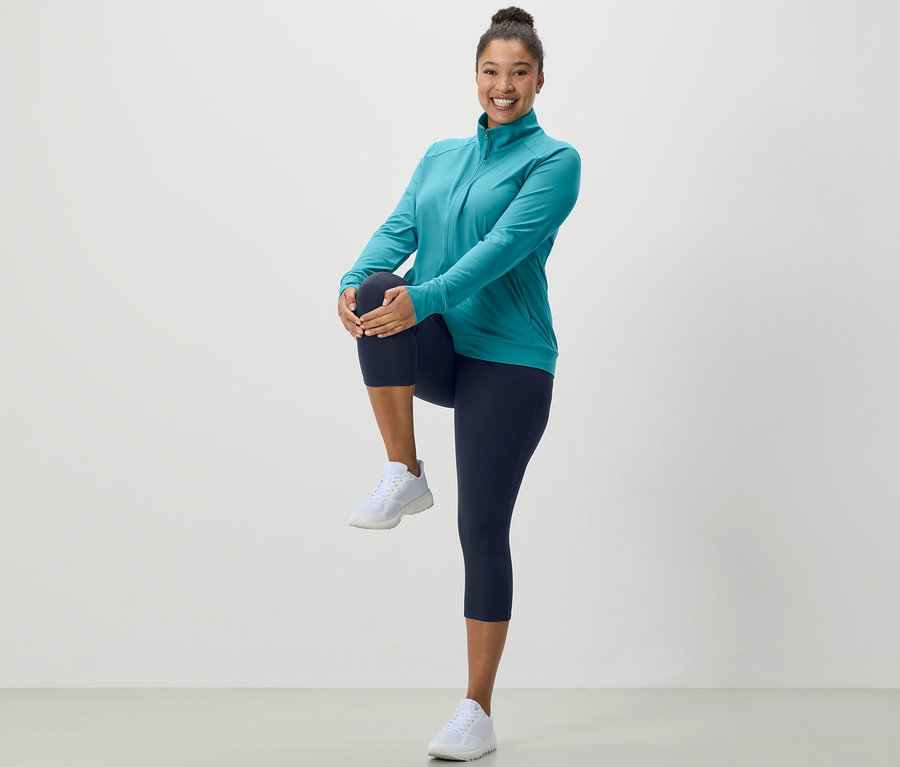 Femme étire jambe en collant de sport 3/4 bleu foncé et veste fonctionnelle turquoise.