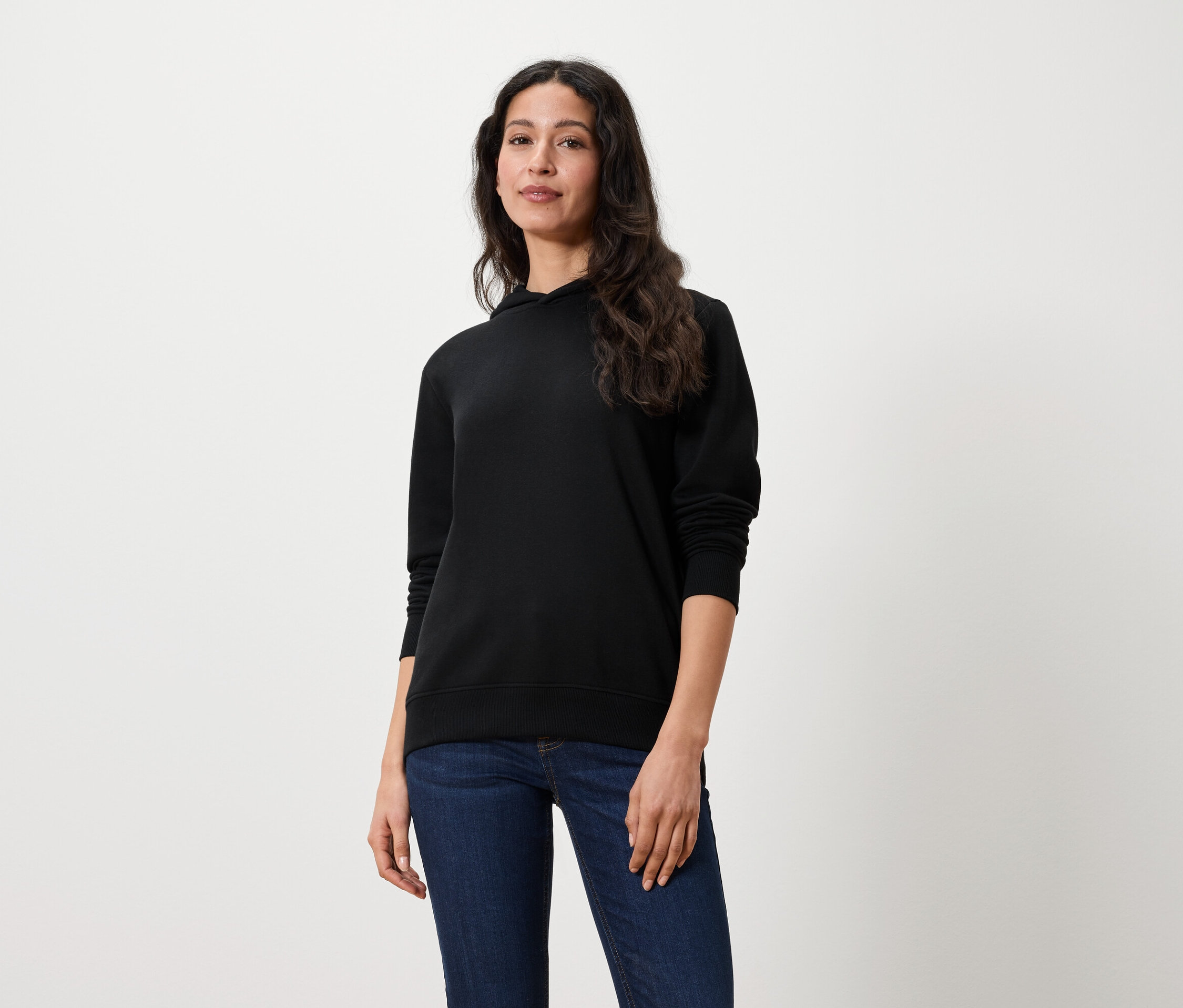 Une femme aux longs cheveux bruns porte un sweat à capuche noir et un jean.