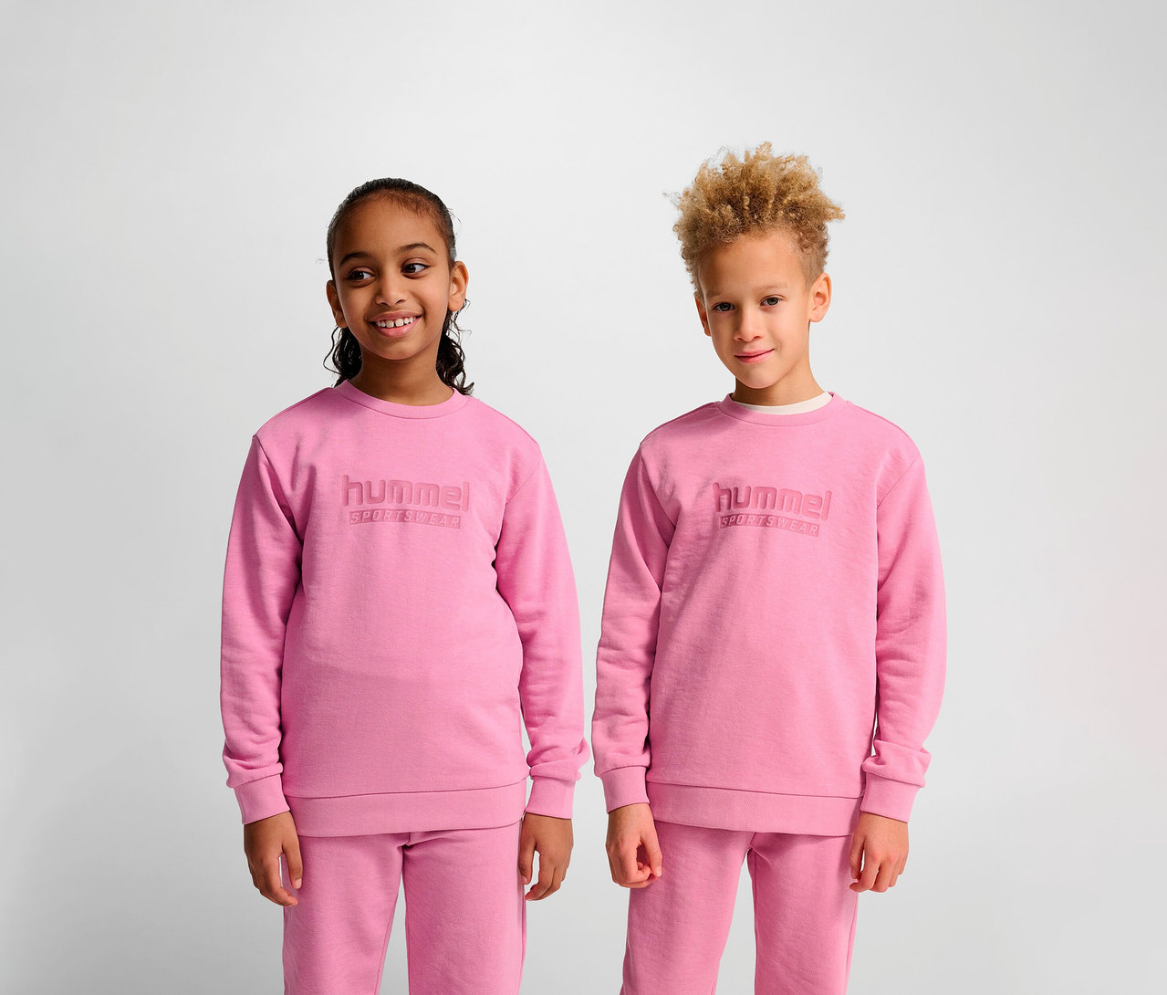 Deux enfants se tiennent côte à côte et portent des sweat-shirts roses Hummel HMLJR Base.