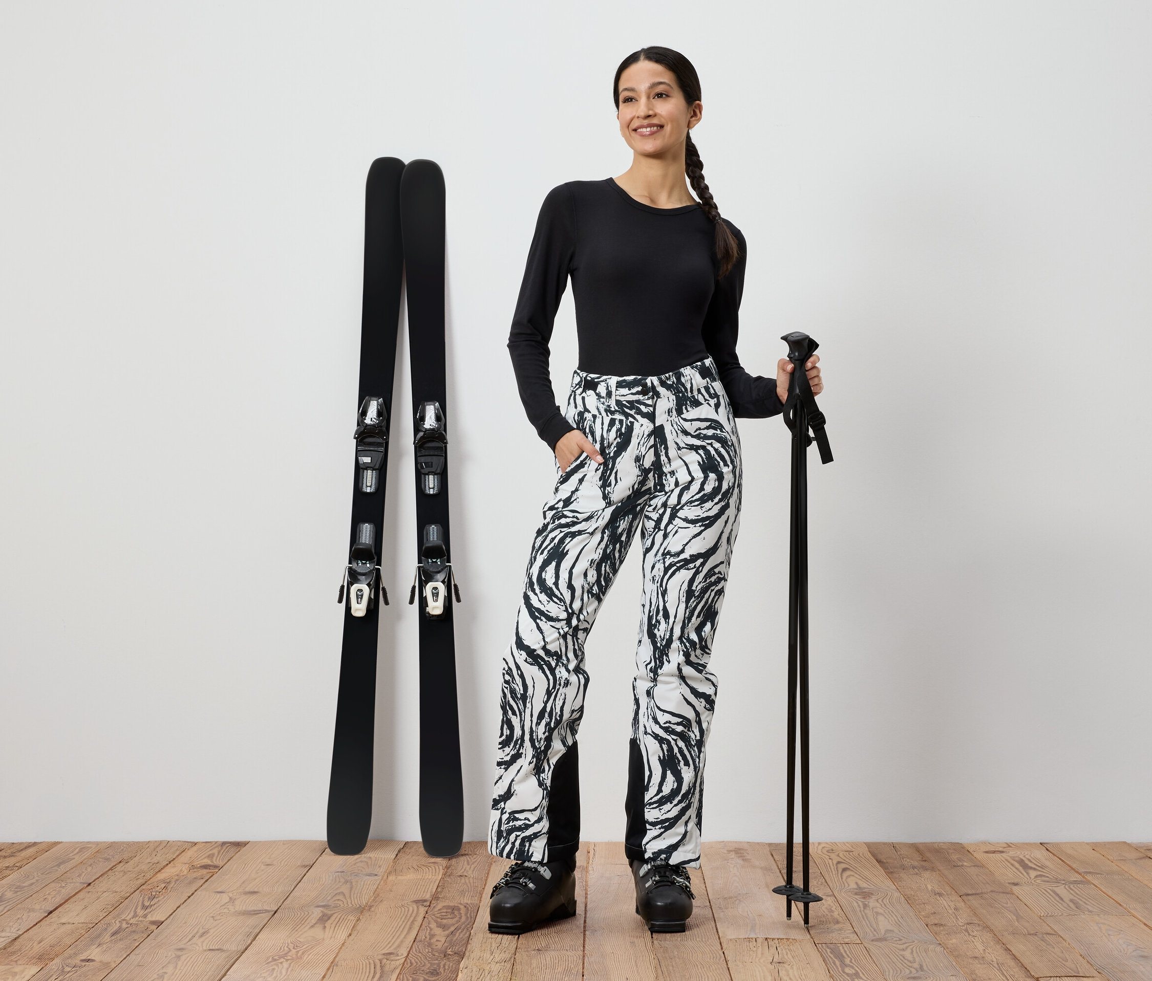 Une femme en haut noir et pantalon à motifs se tient à côté de skis et tient des bâtons de ski.