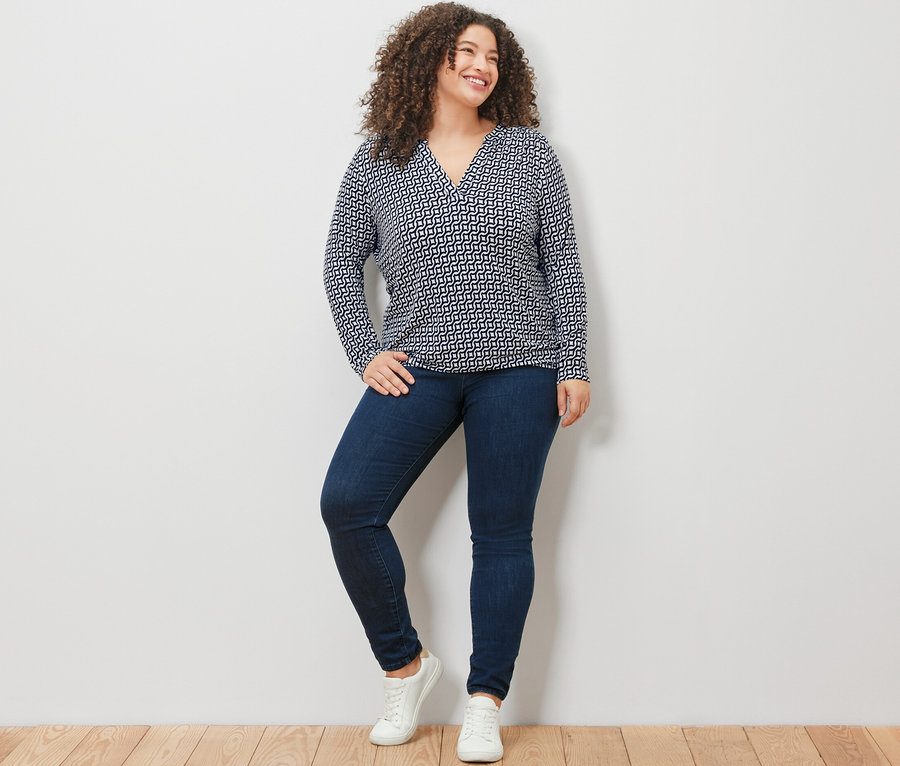 Une femme debout devant un mur blanc porte une blouse imprimée bleu marine, un jean et des baskets blanches.