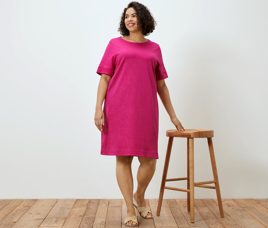 Une femme pose à côté d'un tabouret en bois et porte une robe en jersey rose.