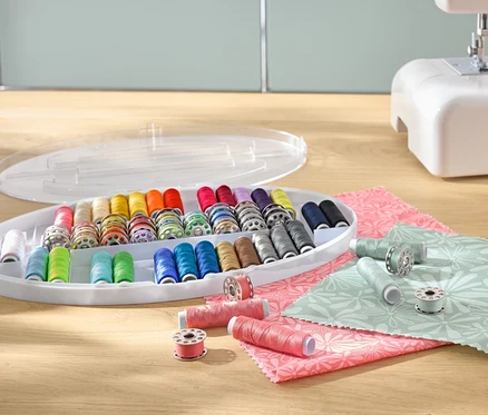 Un kit de fil à coudre avec des bobines de fil colorées dans un organiseur en plastique, à côté d'une machine à coudre et de tissus.