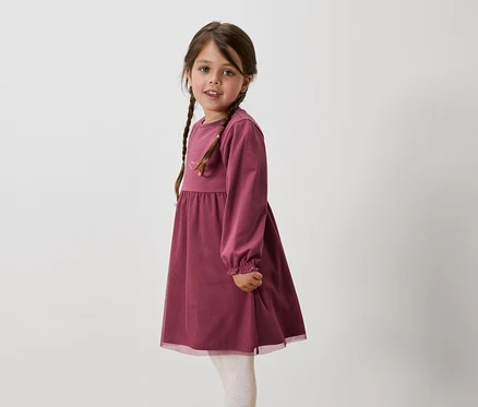 Une fillette aux cheveux bruns tressés porte une robe bordeaux à manches longues et des leggings blancs.