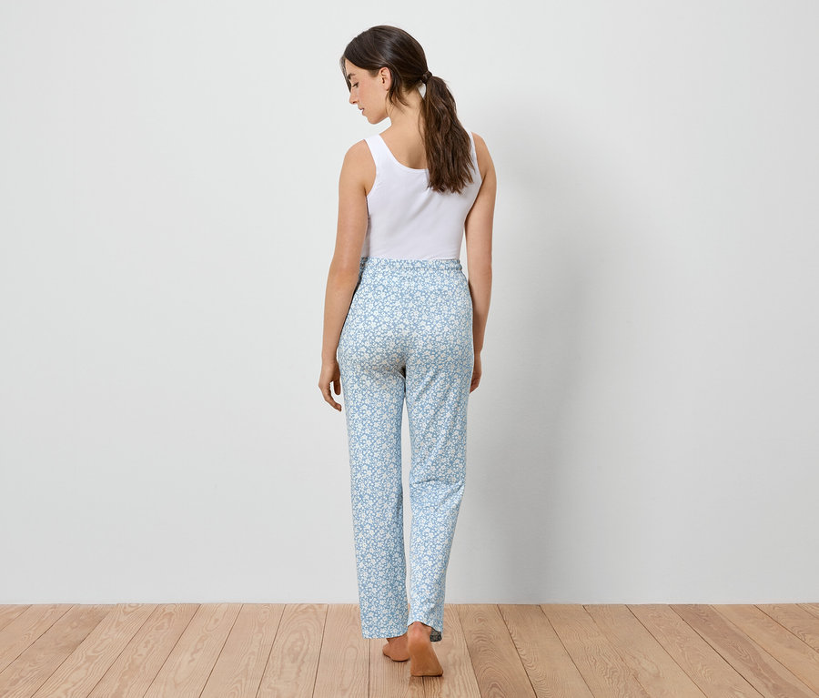 Une femme vue de dos, vêtue d'un débardeur blanc et d'un pantalon bleu clair à motifs floraux, se tient sur un plancher en bois.