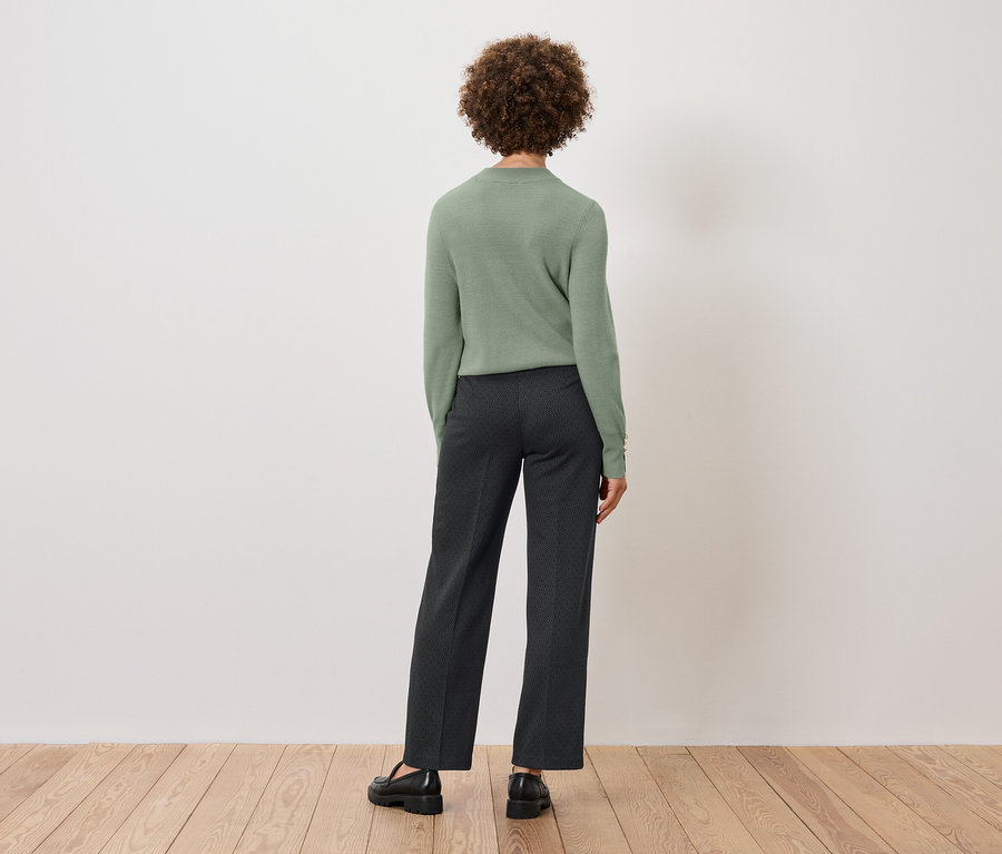 Vue de dos d'une femme aux cheveux bouclés portant un pull vert et un pantalon foncé, debout sur un plancher en bois devant un mur blanc.
