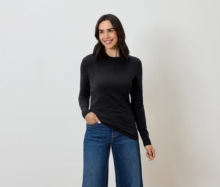 Une femme aux cheveux bruns pose en t-shirt noir à manches longues et en jean bleu.
