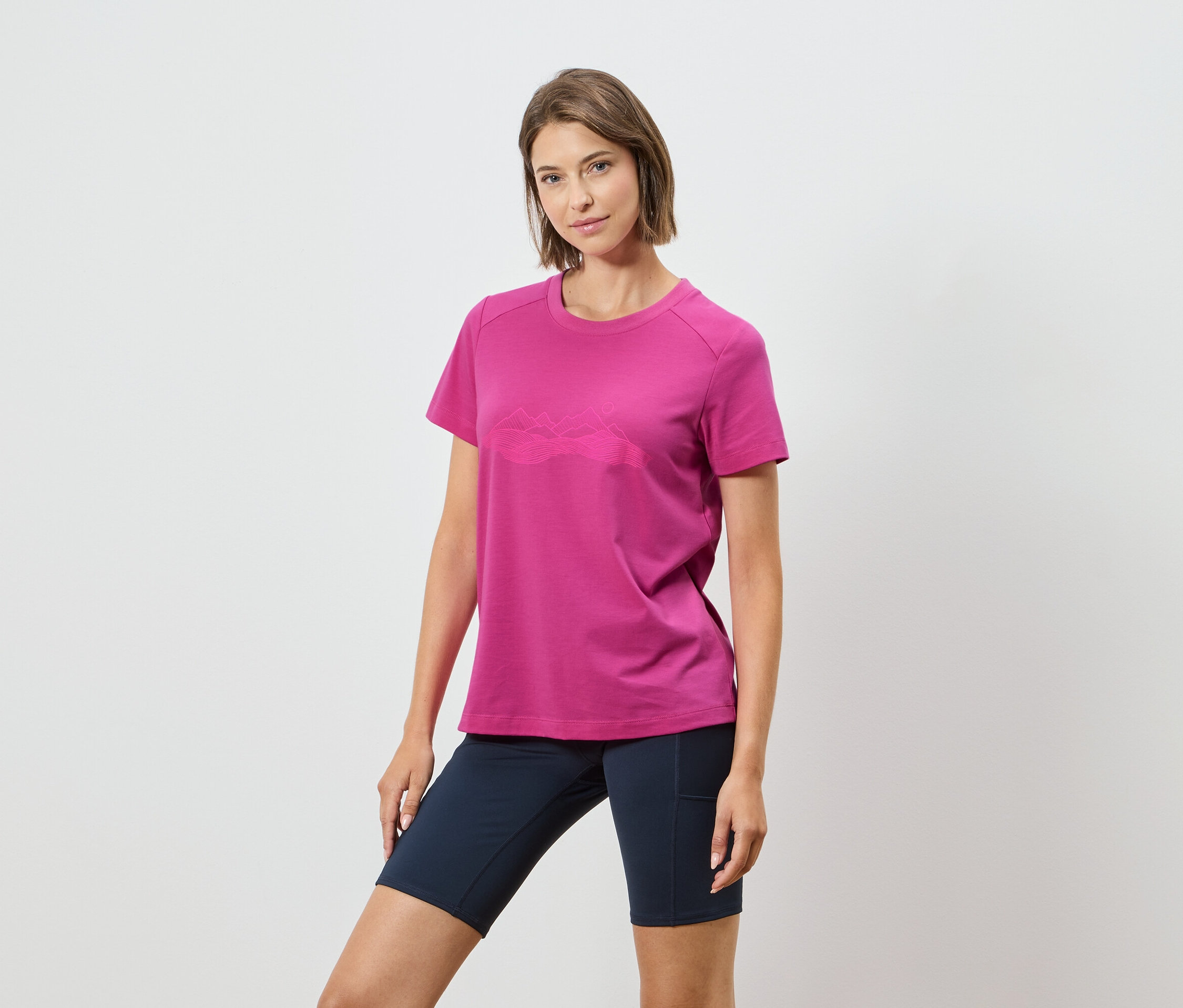Une femme porte un t-shirt rose avec un motif de chaîne de montagnes et un short bleu marine sur un fond blanc.