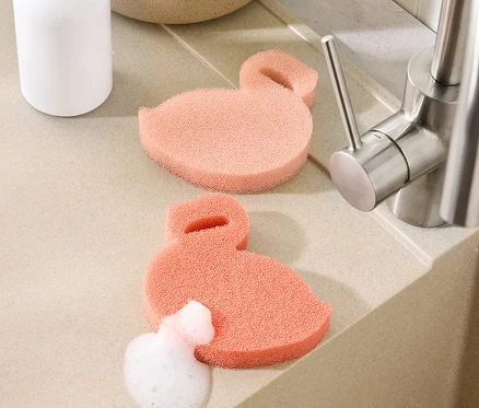 Deux éponges en silicone en forme de flamant rose sont posées sur le plan de travail près d'un évier, l'une d'elles est recouverte de mousse.