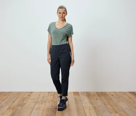Une femme se tient devant un mur blanc sur un plancher en bois. Elle porte un t-shirt vert et un pantalon noir.