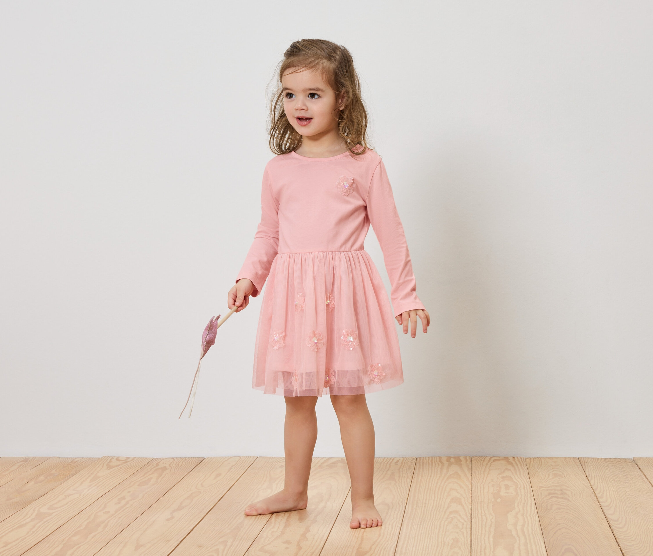 Fillette blonde vêtue d'une robe rose tenant un objet violet à la main.