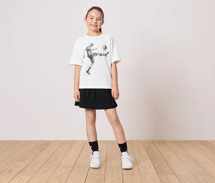 Une jeune fille porte un t-shirt blanc avec un motif de joueur de football, une jupe noire, des chaussettes et des chaussures blanches.