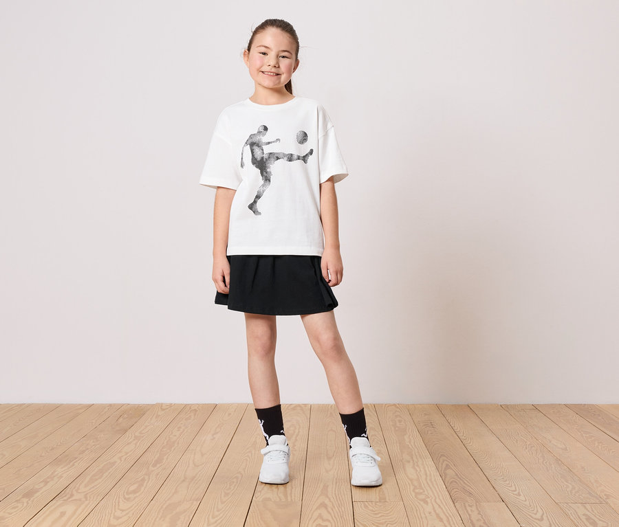 Une jeune fille porte un t-shirt blanc avec un motif de joueur de football, une jupe noire, des chaussettes et des chaussures blanches.