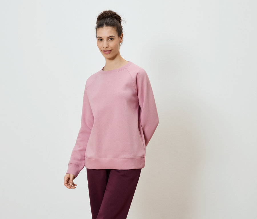 Une femme portant un sweat-shirt rose et un pantalon bordeaux sur un fond blanc.