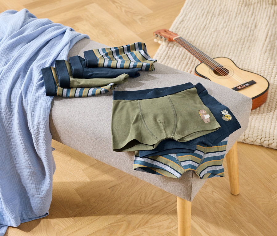 Sur un banc, une couverture bleue et plusieurs boxers verts, bleu marine et à rayures. Une petite guitare est posée à côté.
