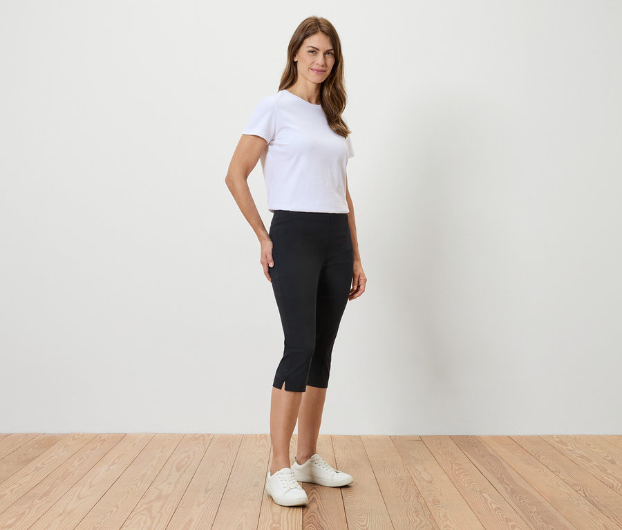 Une femme se tient devant un mur blanc, portant un t-shirt blanc, un pantacourt noir et des baskets blanches.
