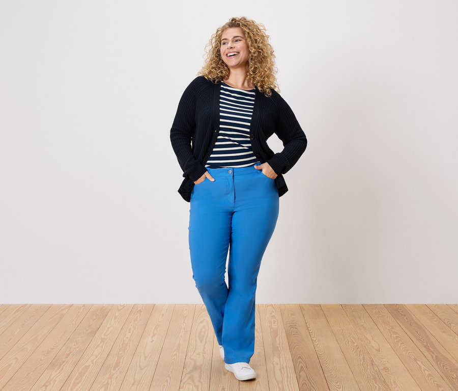 Une femme aux cheveux bouclés porte un cardigan bleu marine, une chemise rayée et un pantalon bleu.
