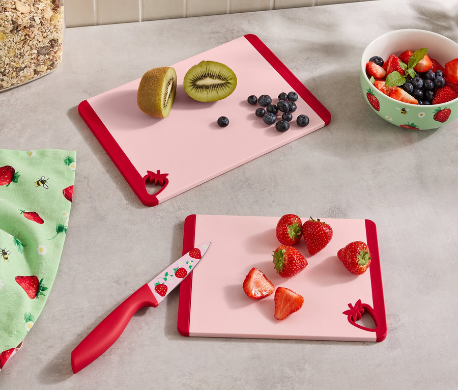 Sur un comptoir de cuisine, il y a deux planches à découper roses avec des bords rouges, un récipient de céréales, un bol de baies, un torchon et un couteau.