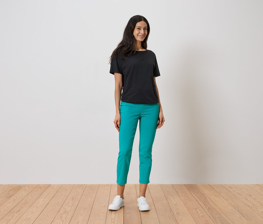 Une femme debout portant un t-shirt noir, un pantalon turquoise et des baskets blanches.