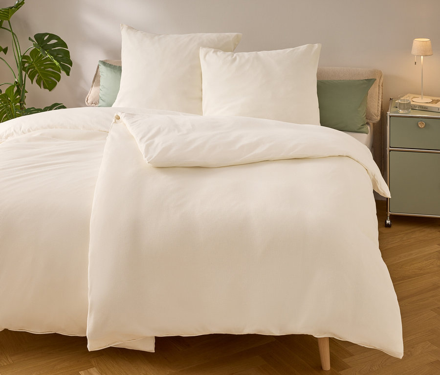 Lit avec des draps et des oreillers beige clair. Une lampe et un livre sont posés sur la table de chevet. Une plante verte est à gauche du lit.