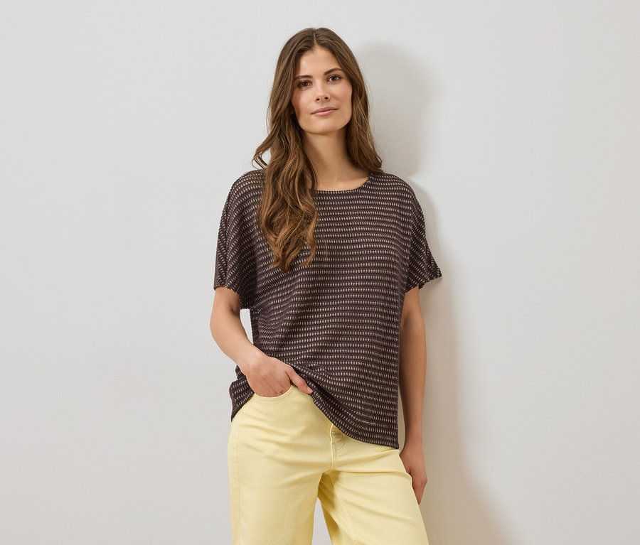 Une femme portant un haut rayé marron à manches courtes et un pantalon jaune, une main dans la poche, devant un mur blanc.