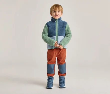 Un garçon blond se tient sur un fond clair, vêtu d'une veste en polaire bleue, verte et blanche, d'un pantalon orange et de bottes bleues.