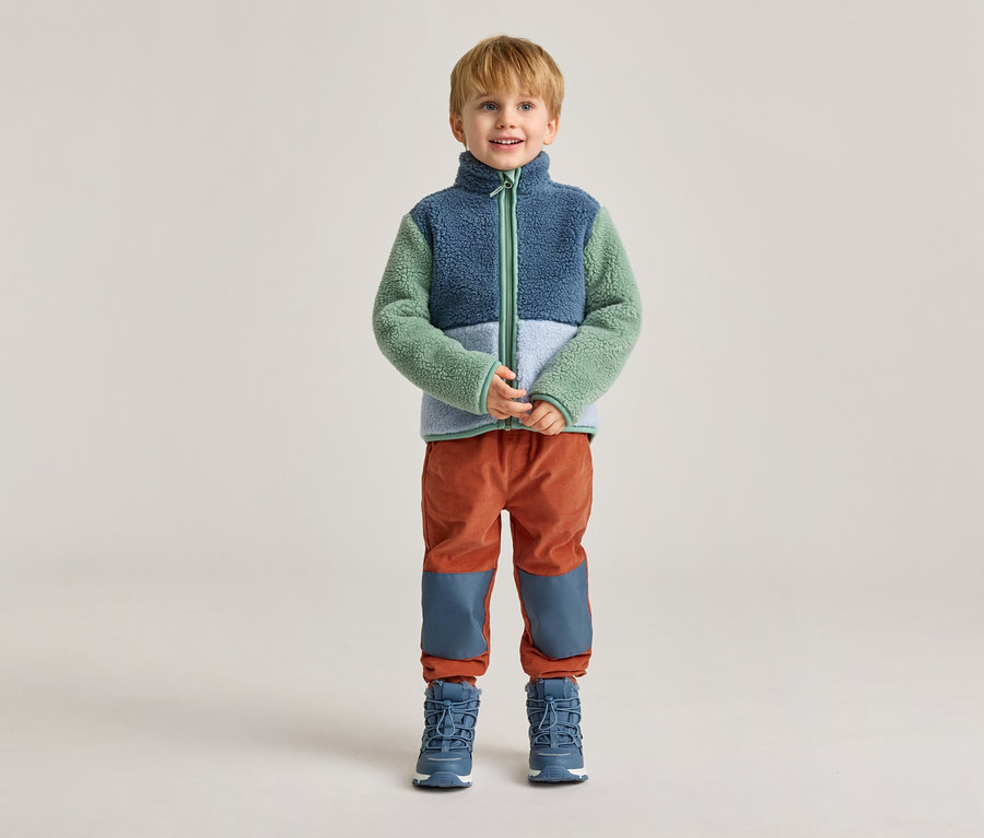 Un garçon blond se tient sur un fond clair, vêtu d'une veste en polaire bleue, verte et blanche, d'un pantalon orange et de bottes bleues.